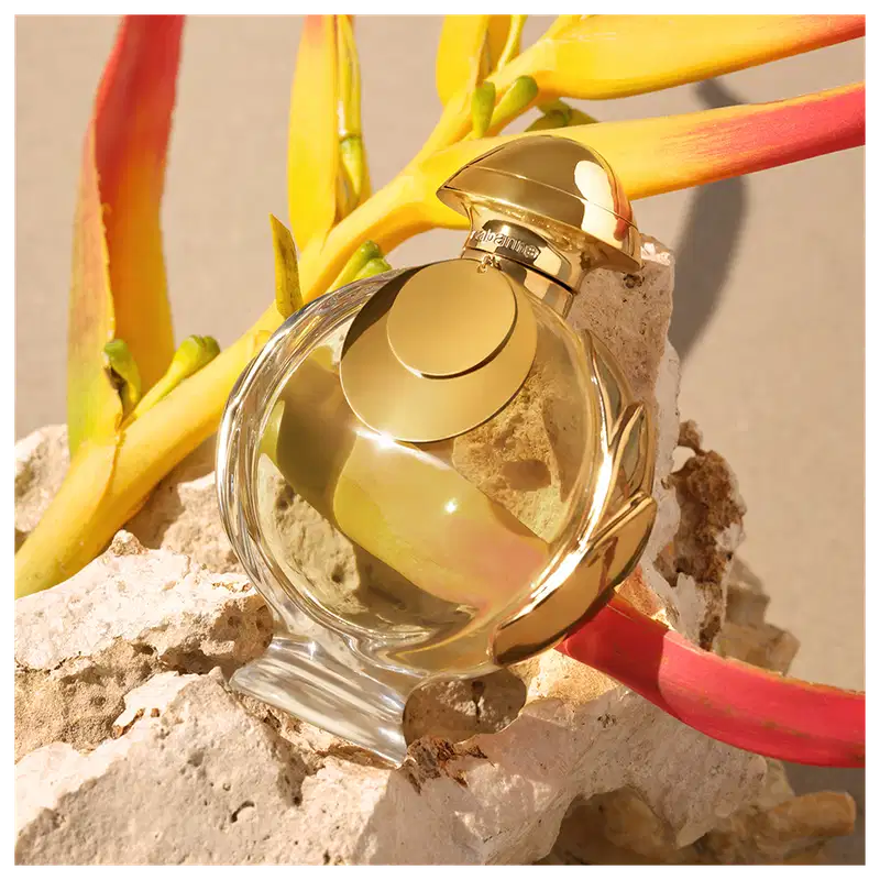 Recipiente de item de perfumaria dourado em primeiro plano, com flores amarelas e vermelhas ao redor, sobre pedras em um ambiente natural.
