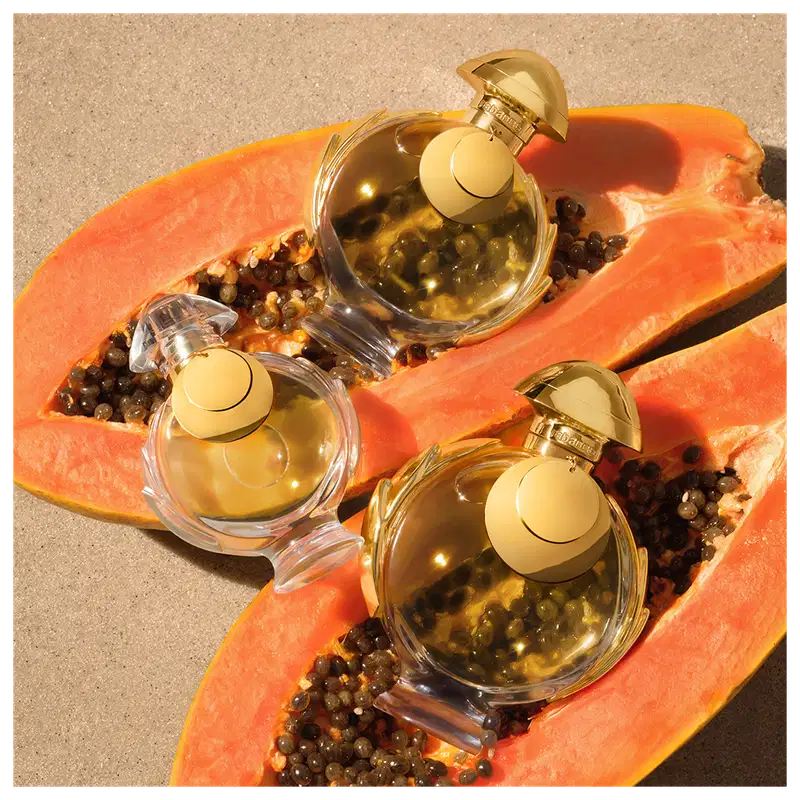 Três frascos de item de perfumaria dourados sobre fatias de mamão papaia laranja em um fundo bege. Os frascos têm tampas douradas e contêm um líquido dourado. 