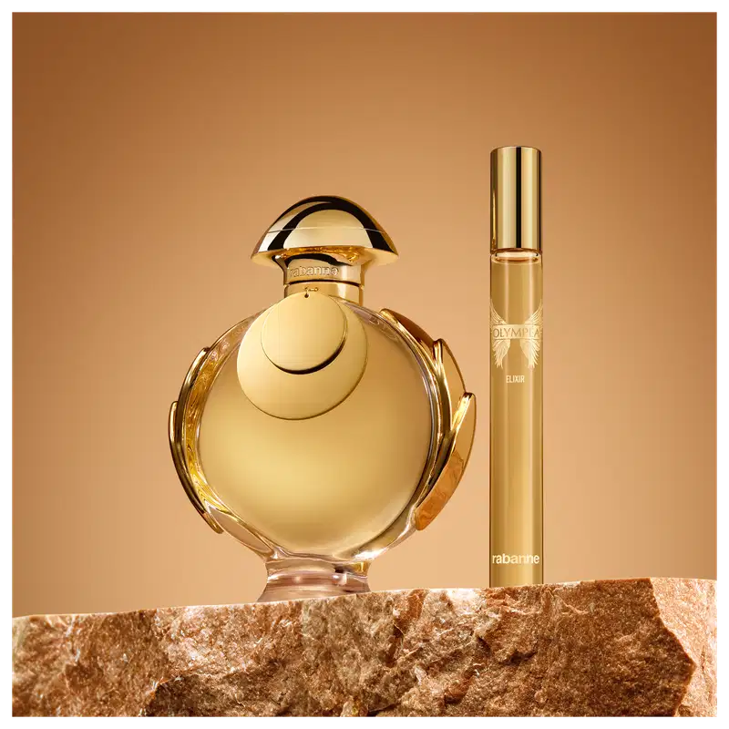 Ambiente de exibição de item de perfumaria da marca Olympea, com frasco de vidro dourado e tampa arredondada, acompanhado de um frasco menor, sobre superfície de pedra marrom em fundo laranja.