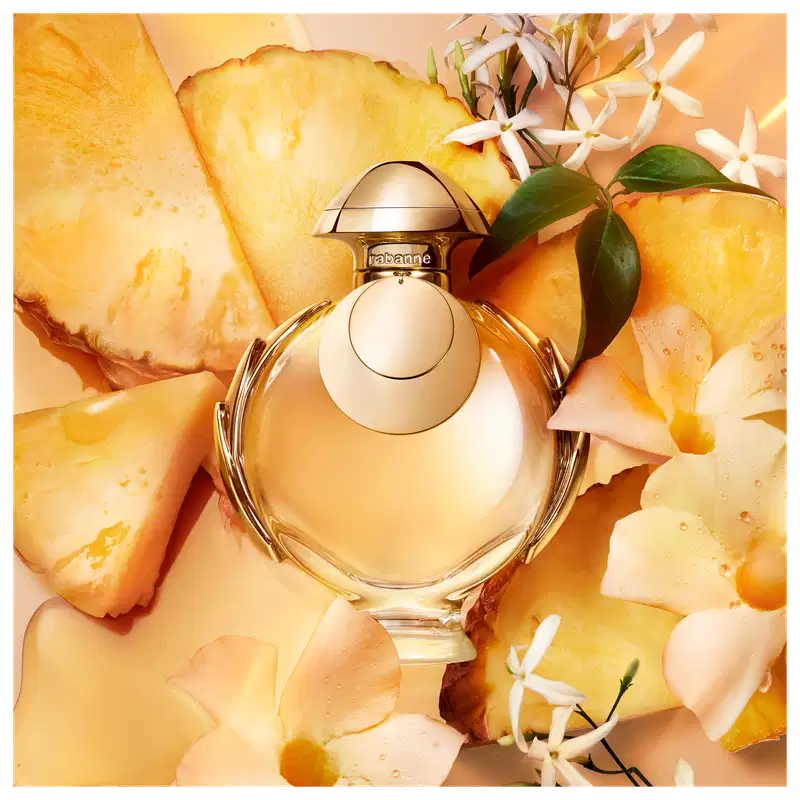 Recipiente de item de perfumaria Trabanne em primeiro plano, rodeado por fatias de abacaxi amarelo-dourado e flores brancas. 
