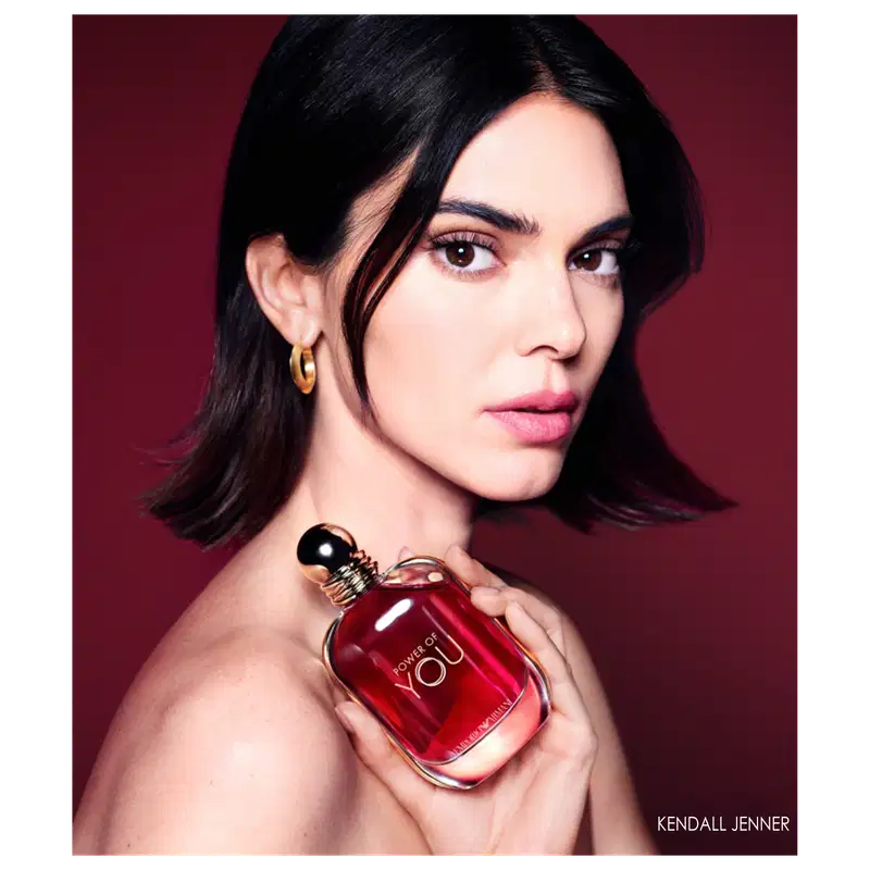 Mulher com cabelos escuros, modelo Kendall Jenner, e olhar intenso segurando o perfume. Ambiente de retrato de moda com iluminação dramática e cores contrastantes.