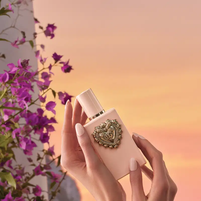 Recipiente de item de perfumaria com design ornamentado em forma de coração, posicionado em ambiente externo com vegetação florida e céu ao fundo em tons de laranja e rosa.