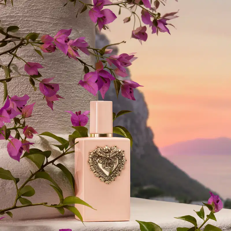Recipiente de item de perfumaria com design ornamentado em forma de coração, posicionado em ambiente externo com vegetação florida e céu ao fundo em tons de laranja e rosa.