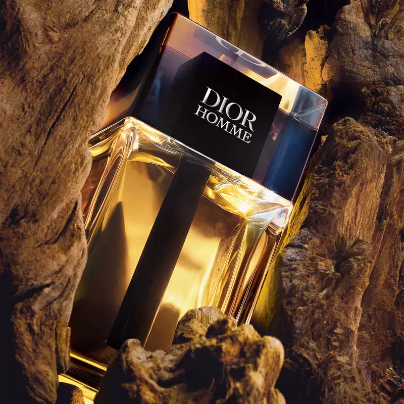 Embalagem do perfume da marca Dior em primeiro plano, sobre superfície de madeira rústica com pedras ao redor, em ambiente escuro e iluminado.