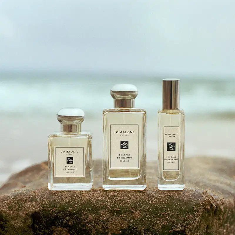 Três frascos de perfume da marca Jo Malone London, com a fragrância Sea Salt & Bergamot, em diferentes volumetrias, dispostos sobre uma rocha em um ambiente natural, com o mar ao fundo, sugerindo um cenário costeiro e relaxante.