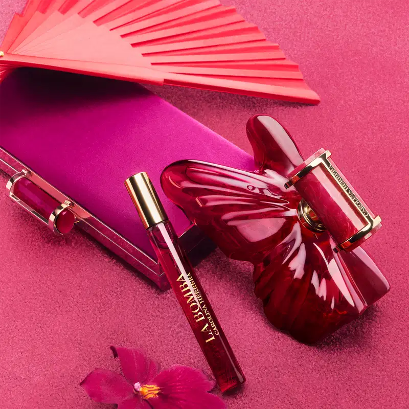 Conjunto de produtos de beleza da marca Carolina Herrera, incluindo um frasco de perfume com tampa dourada e um travel size, todos com embalagens em tons de rosa intenso e detalhes dourados.