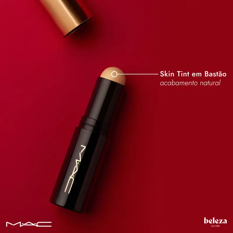 Bastão de maquiagem M·A·C com tampa dourada e corpo preto, texto "Skin Tint em Bastão" e "acabamento natural" visível na embalagem, sobre fundo vermelho.