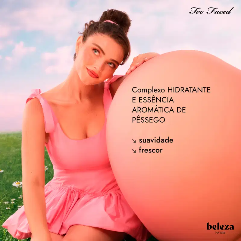 Em campo gramado e florido com céu ao fundo, jovem de vestido rosa encosta-se em grande esfera cor de pêssego. A marca "Too Faced" está no topo. Na esfera lê-se "Complexo HIDRATANTE E ESSÊNCIA AROMÁTICA DE PÊSSEGO", "suavidade", "frescor"