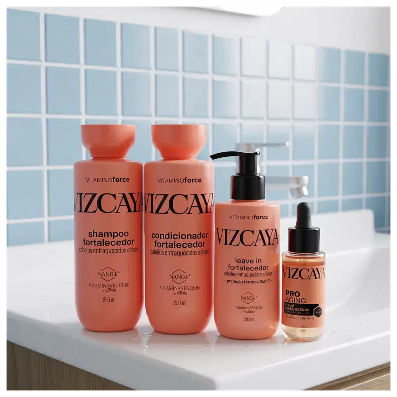 A coleção de produtos Vizcaya incluindo shampoo, condicionador, leave-in e sérum para cabelos, em embalagens de plástico laranja com rótulos em preto, dispostos sobre uma superfície branca com azulejos ao fundo.