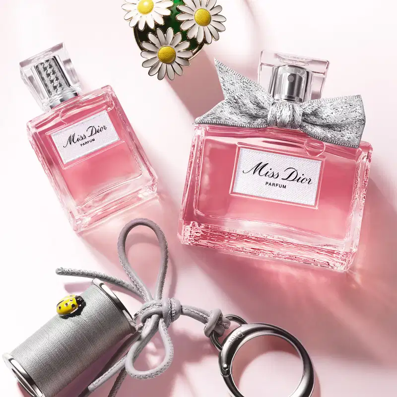 Dois frascos Miss Dior Parfum rosa em superfície rosa claro, acompanhados de margaridas brancas com centro amarelo, chaveiro cinza com laço prateado e joaninha amarela.