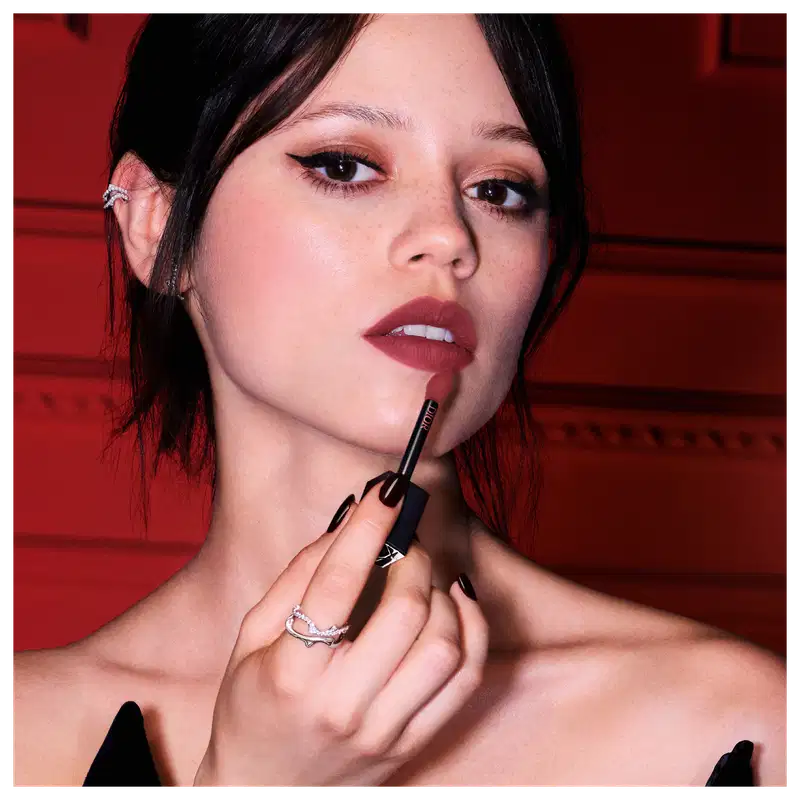Atriz Jenna Ortega, mulher branca com cabelos curtos escuros, segura aplicador do batom líquido enquanto usa em seus lábios um batom vermelho.