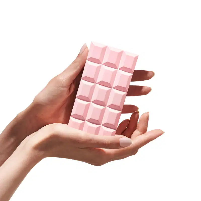 Mão segurando a embalagem de perfume parecendo um Painel de parede 3D rosa pastel com design geométrico, composto por 12 módulos quadrados em relevo, criando um efeito visual tridimensional elegante e moderno.