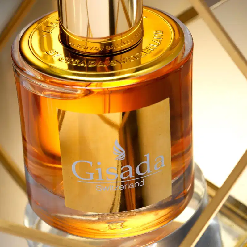Perfume Gisada Switzerland em frasco de vidro transparente com líquido âmbar. Tampa dourada com detalhes em ouro. Rótulo retangular dourado com logo e nome da marca em letras prateadas. Design elegante e luxuoso.