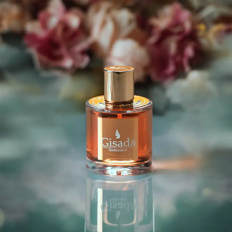 Frasco de perfume Gisada com líquido âmbar em ambiente floral. Flores rosas e brancas desfocadas ao fundo criam cenário elegante. Superfície reflexiva realça o tom dourado do frasco com tampa cilíndrica. Contexto sofisticado para produto de luxo.