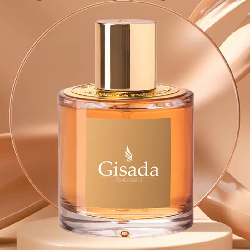 Perfume Gisada em frasco cilíndrico de vidro com líquido âmbar. Tampa dourada cilíndrica. Rótulo bege com logo branco e texto "Gisada Switzerland". Apresentação luxuosa em fundo neutro com efeitos de luz.