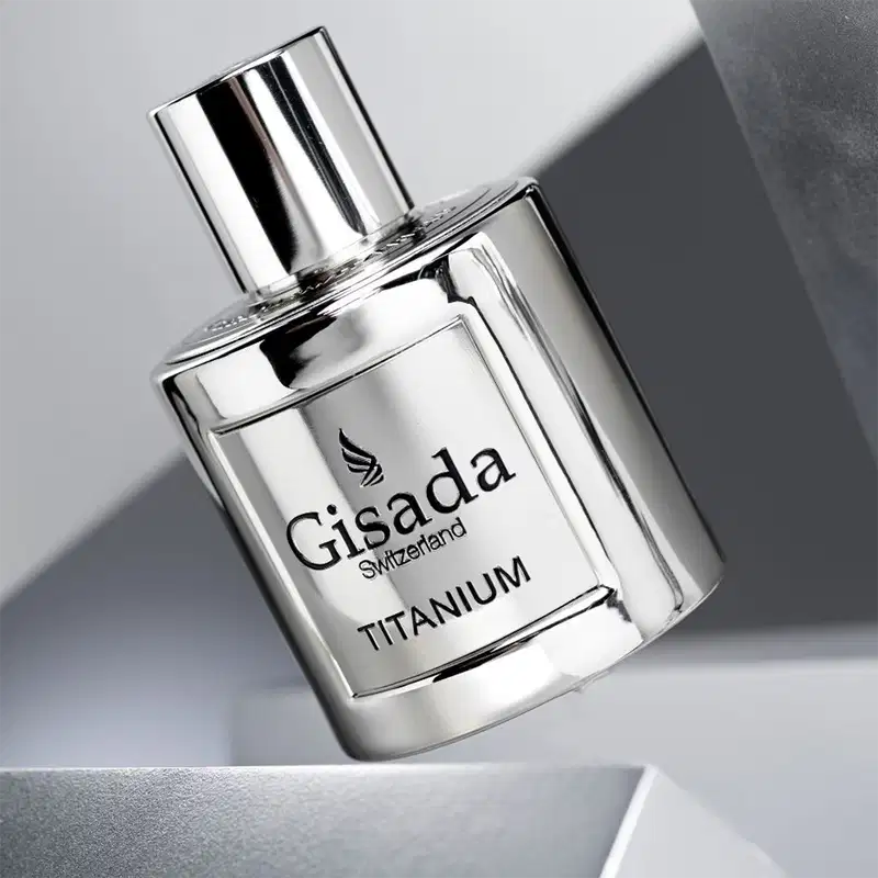Perfume Gisada Titanium em frasco de vidro transparente com tampa prateada cilíndrica. Embalagem branca e prata com logo alado da marca e texto "Gisada Switzerland TITANIUM" em preto. Design minimalista e elegante.