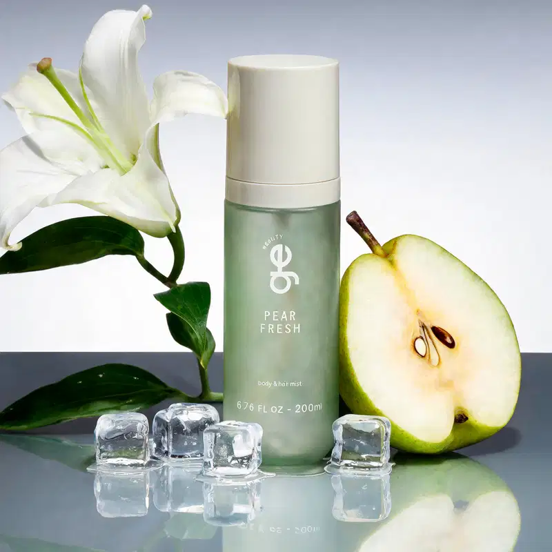 Ambiente sofisticado e minimalista com fundo cinzento. Produto GE Beauty Pear Fresh em frasco verde-água com tampa branca, cercado por pera fresca cortada, flor branca de lírio, folhas verdes e cubos de gelo. Composição que evoca frescor.