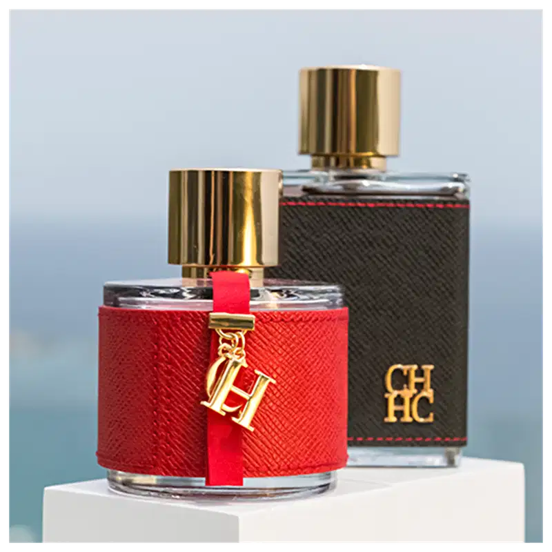 Dois frascos de perfume da marca Carolina Herrera, um vermelho e outro preto, com detalhes dourados e logotipo da marca. O frasco vermelho possui uma fita vermelha e um pingente dourado com as iniciais "CH".