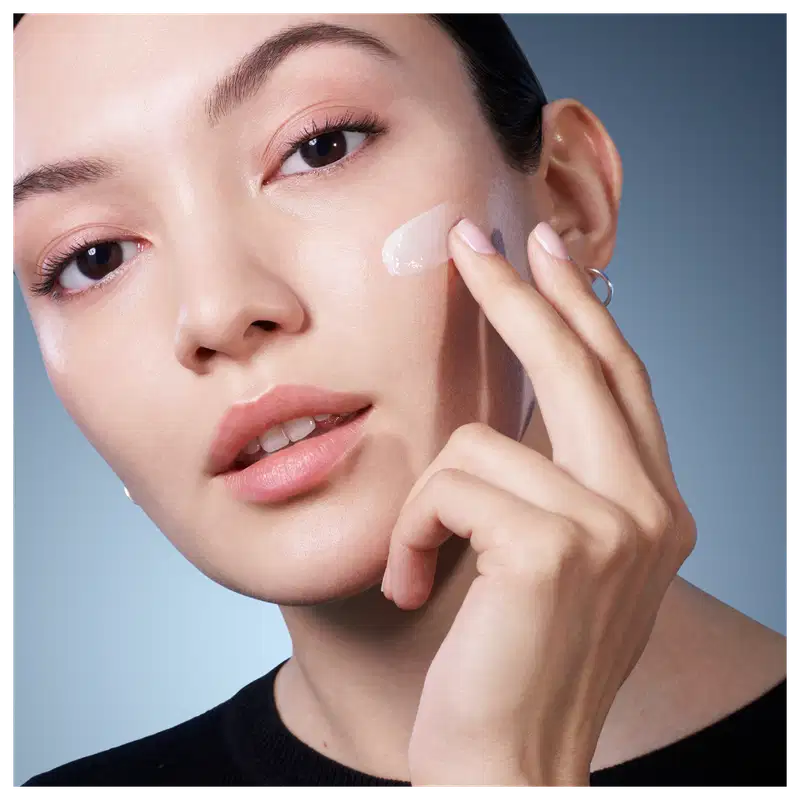 Retrato de uma mulher aplicando um produto de cuidados com a pele da marca Lancôme. Fundo azul claro, iluminação suave, expressão serena e concentrada.
