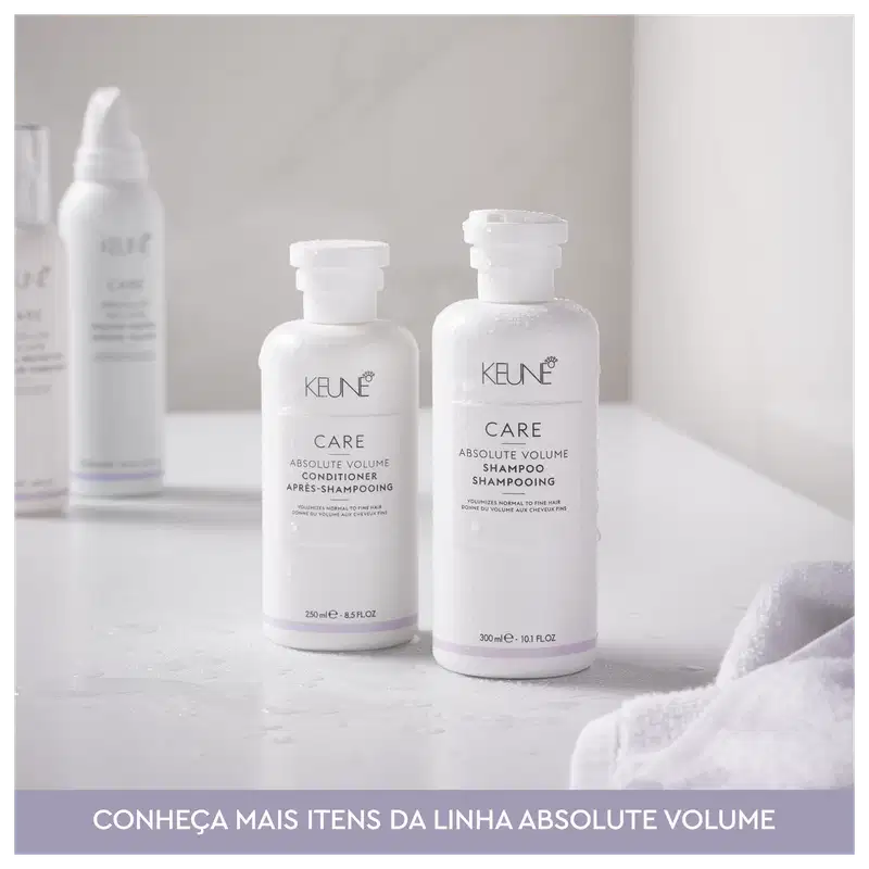 Dois frascos brancos da linha Absolute Volume da Keune, um contendo condicionador e outro shampoo, com rótulos e logotipos da marca. Os produtos são apresentados em embalagens de plástico branco com detalhes em roxo.