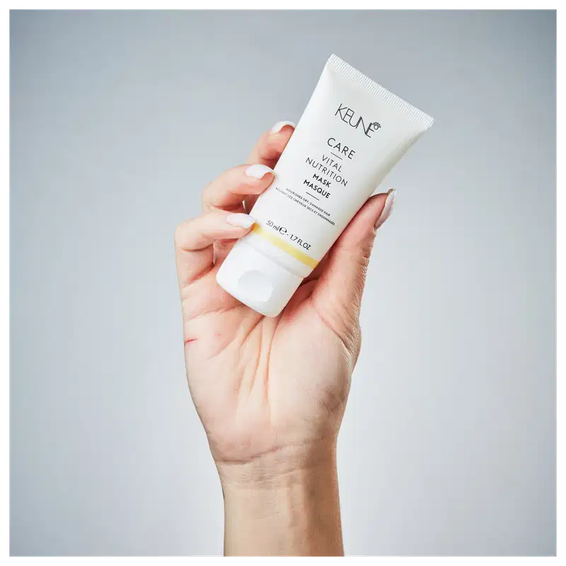 Mão segurando um tubo de máscara facial da marca Keune, em um ambiente minimalista e monocromático, sugerindo um contexto de cuidados com a pele e beleza.