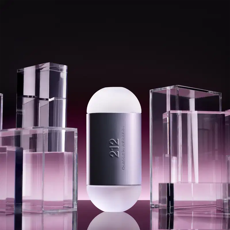 Frasco de perfume Carolina Herrera 212 em formato oval, com superfície prateada e detalhes em branco. O rótulo exibe o nome da fragrância em letras grandes.