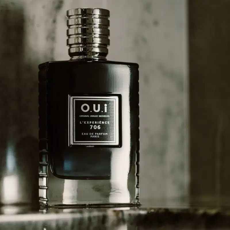 Um item de perfumaria preto com rótulo branco e texto em francês, posicionado em um ambiente escuro e sombrio, sugerindo um contexto elegante e sofisticado para seu uso.