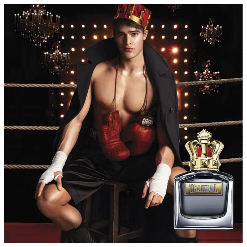 O ambiente retratado é um ringue de boxe iluminado por luzes brilhantes, com um homem musculoso vestindo luvas vermelhas e um casaco preto, segurando um frasco de perfume da marca Jean Paul Gaultier, sugerindo uma associação entre o produto e o esporte.