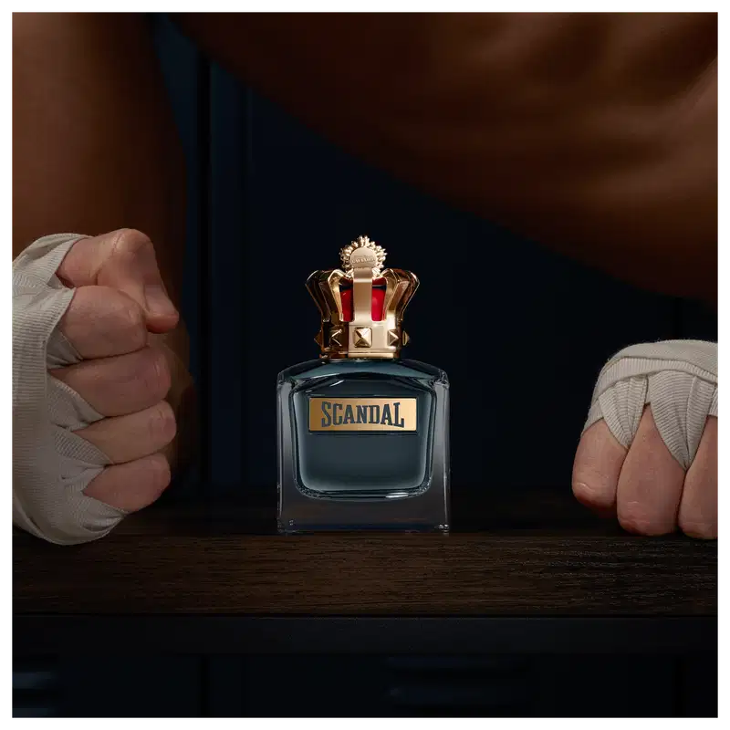 Frasco de perfume Jean Paul Gaultier Scandal, com tampa dourada em formato de coroa e rótulo em fundo azul escuro. Embalagem elegante e sofisticada, com design marcante e distintivo da marca.