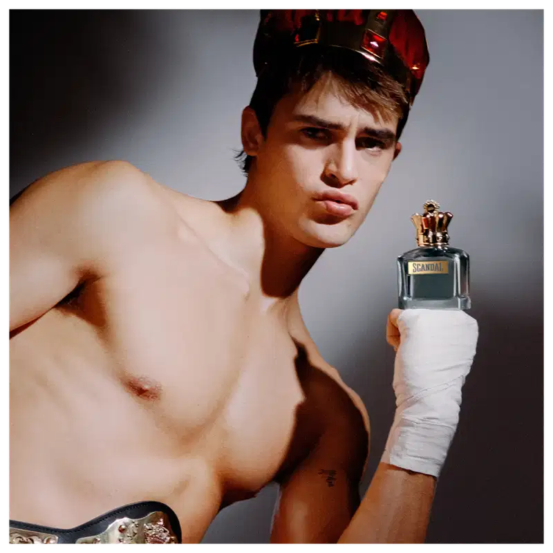 Ambiente escuro e dramático, com modelo masculino segurando um frasco de perfume da marca Jean Paul Gaultier, destacando-se com sua expressão séria e pose desafiadora.