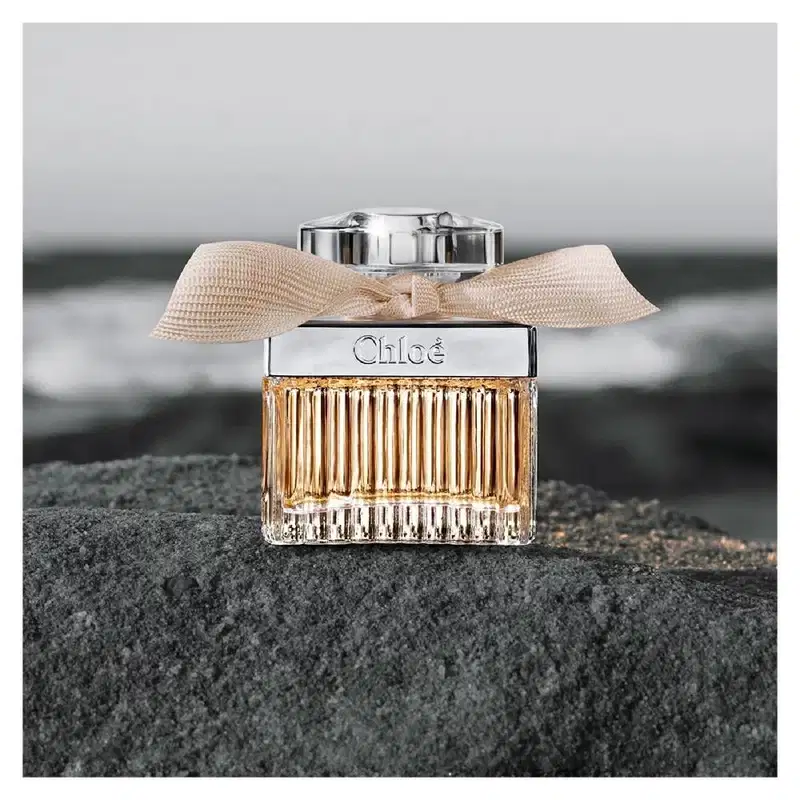 Frasco de perfume Chloé com tampa prateada e laço bege, apresentando o nome da marca em destaque. O design elegante e minimalista transmite sofisticação.