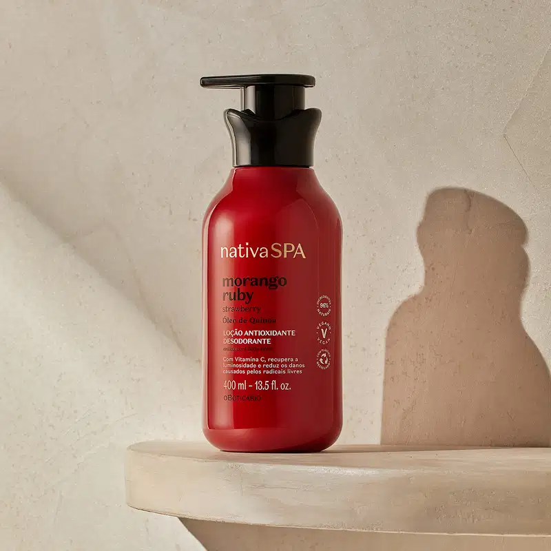 Frasco vermelho com bomba dosadora preta e letras douradas, brancas e pretas de loção antioxidante corporal Nativa SPA Morango Ruby posicionado em superfície bege clara, com sombra geométrica ao fundo e parede texturizada em tons neutros.