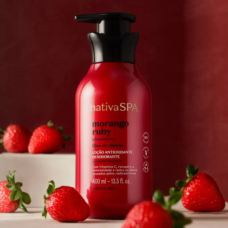 Frasco vermelho com bomba dosadora preta e letras douradas, brancas e pretas de loção antioxidante corporal Nativa SPA Morango Ruby, cercado por morangos frescos sobre superfície branca e fundo vermelho escuro.