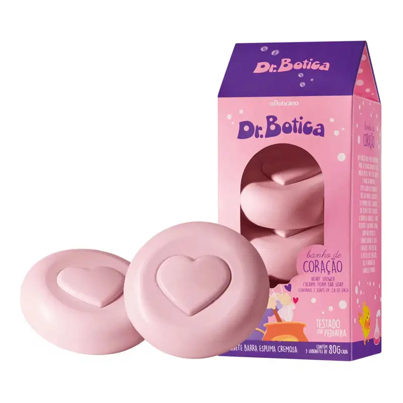 Caixa rosa e azul escuro ao lado de dois sabonetes em barra na cor rosa com coração gravado nos itens da marca Dr. Botica.