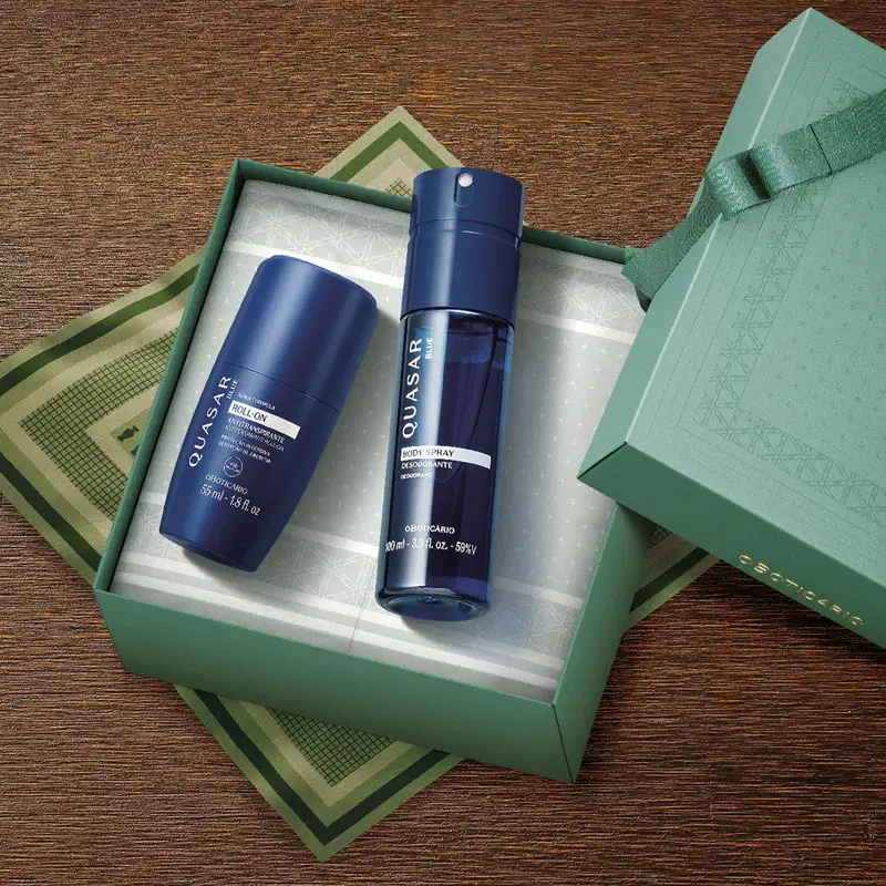 Imagem de uma caixa verde com detalhe dourado contendo dois itens da marca Quasar Blue: um desodorante roll-on e um body spray, ambos em embalagens azuis, sobre superfície de madeira marrom.
