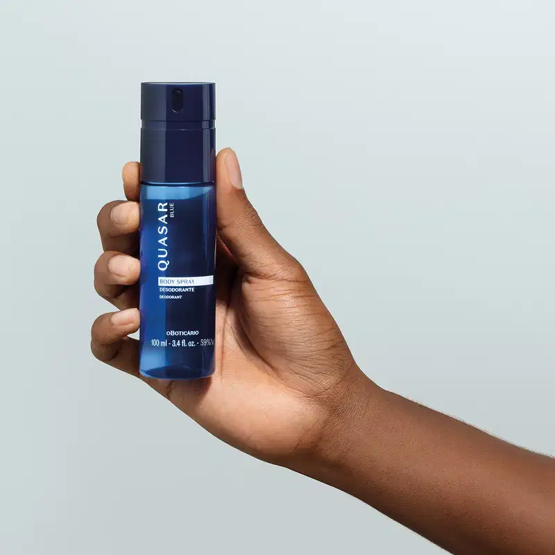 Imagem de embalagem azul escura de body spray Quasar Blue sendo segurada por mão masculina em fundo azul claro.