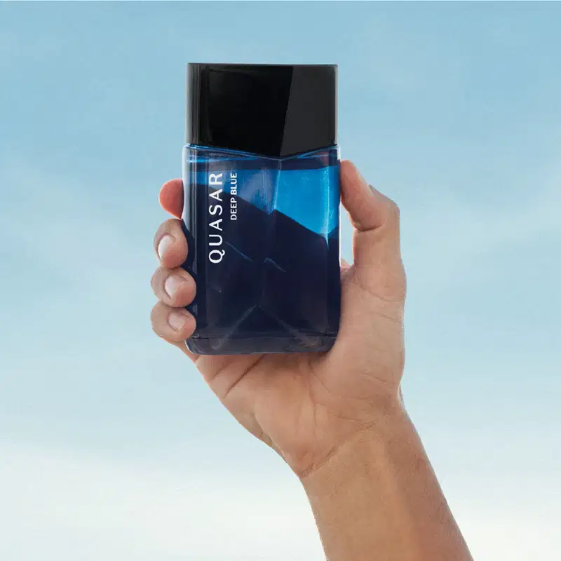 Imagem de frasco de desodorante colônia da marca Quasar Deep Blue em tons de azul, sendo segurado por uma mão em um fundo azul claro.