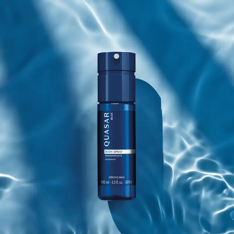 Imagem de um frasco de body spray da marca Quasar Blue em azul escuro, com névoa e reflexos em fundo azul claro e desfocado.