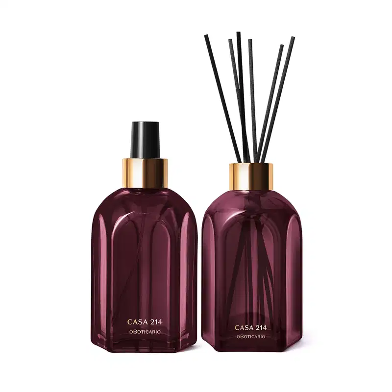 A imagem mostra dois vidros retangulares roxos, com rótulo em dourado. Um deles é o aromatizador de ambientes com válvula spray preta e o outro é um difusor com tampa dourada que acompanha varetas de madeira. Ambos são a linha Casa 214 Figo Magnífico.