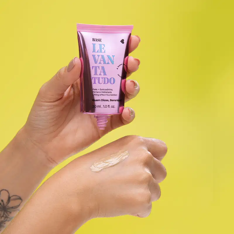 Uma mão feminina segura uma bisnaga rosa metalizada com tampa preta e letras azuis e pretas de base líquida Levanta Tudo da marca Quem Disse, Berenice?, colocando uma pequena quantidade do produto na outra mão, sobre fundo amarelo.