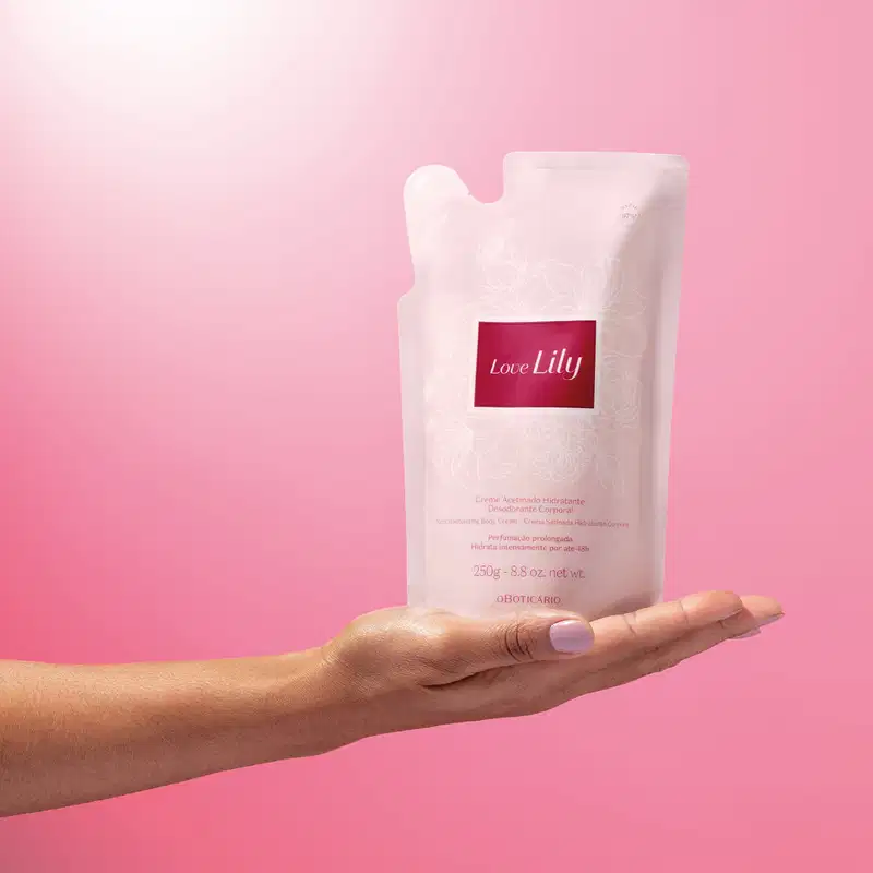 Mão feminina segurando um refil de creme acetinado desodorante hidratante corporal da marca Love Lily em sachê na cor bege com detalhes dourados e rótulo marrom claro. O fundo da imagem é em tons de rosa.