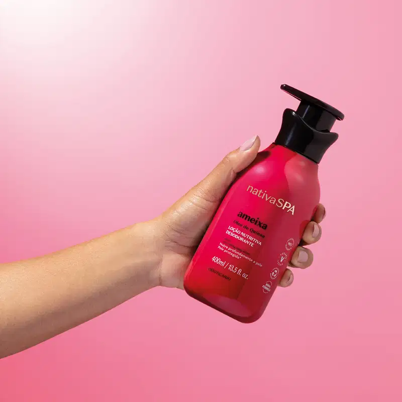 Mão segurando um frasco rosa vibrante com tampa dosadora preta e letras douradas, brancas e pretas de loção hidratante corporal Nativa SPA Ameixa sobre fundo rosa claro.