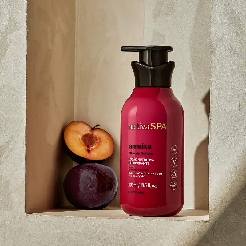 Frasco rosa vibrante com tampa dosadora preta e letras douradas, brancas e pretas de loção hidratante corporal Nativa SPA Ameixa. posicionado em nicho bege ao lado de meia ameixa fresca e ameixa inteira.