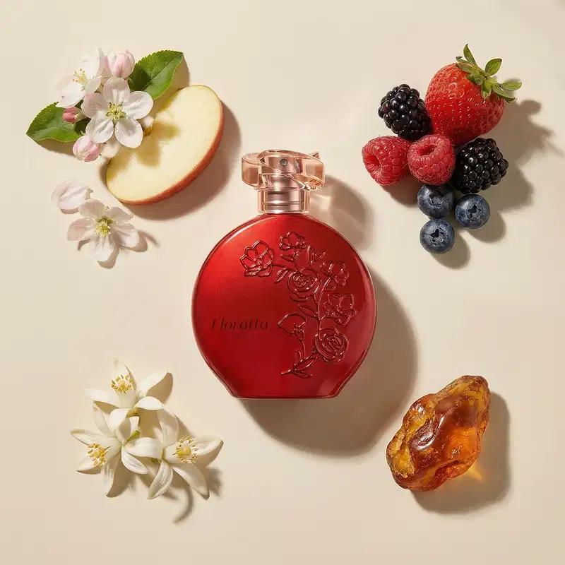 Frasco de item de perfumaria da marca Floratta Red em formato arredondado na cor vermelha, com relevo de flores e do nome da marca cercado por flores brancas, uma pepita de âmbar, frutas vermelhas variadas e uma fatia de maçã.