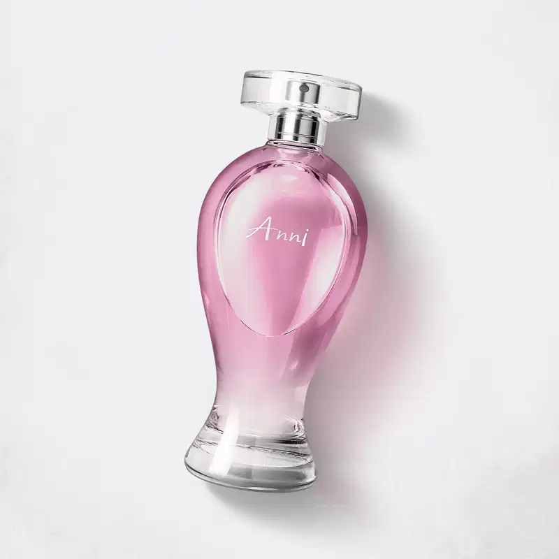 Vidro transparente em formato curvilíneo com líquido rosa, pulverizador prateado e tampa transparente contendo 200ml de desodorante colônia Boticollection Anni em um fundo cinza.