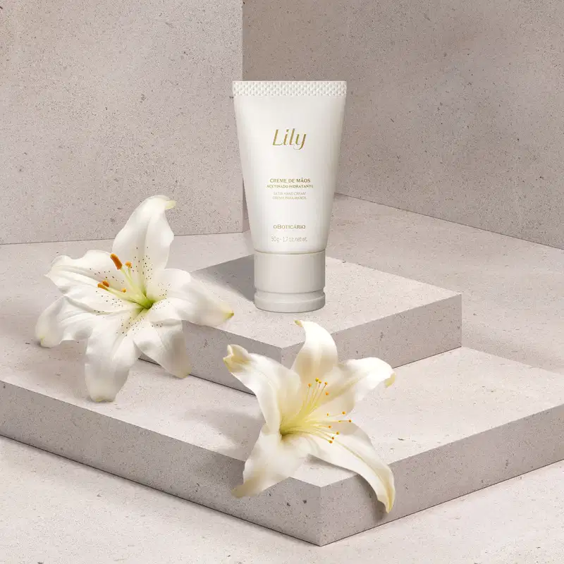 Uma biisnaga branca com tampa bege e letras marrons de creem de mãos acetinado hidratante da marca Lily está em uma plataforma branca, com flores brancas ao redor, em um cenário em tons de cinza.