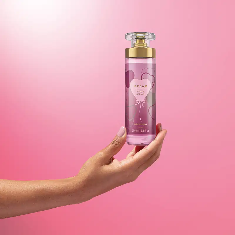 Imagem de uma mão segurando um frasco de body splash Dream Amor no Ar. A embalagem conta com tampa transparente, válvula spray dourada e rotulo em tons de roxo com coração ilustrativo . O cenário traz um fundo em tons de lilás e branco.