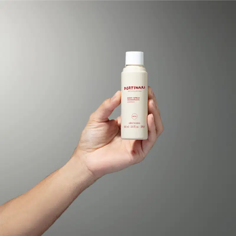 Mão feminina segurando um refil de body spray Boticollection Portinari em frasco na cor branca com letras marrons e tampa branca sobre fundo cinza.