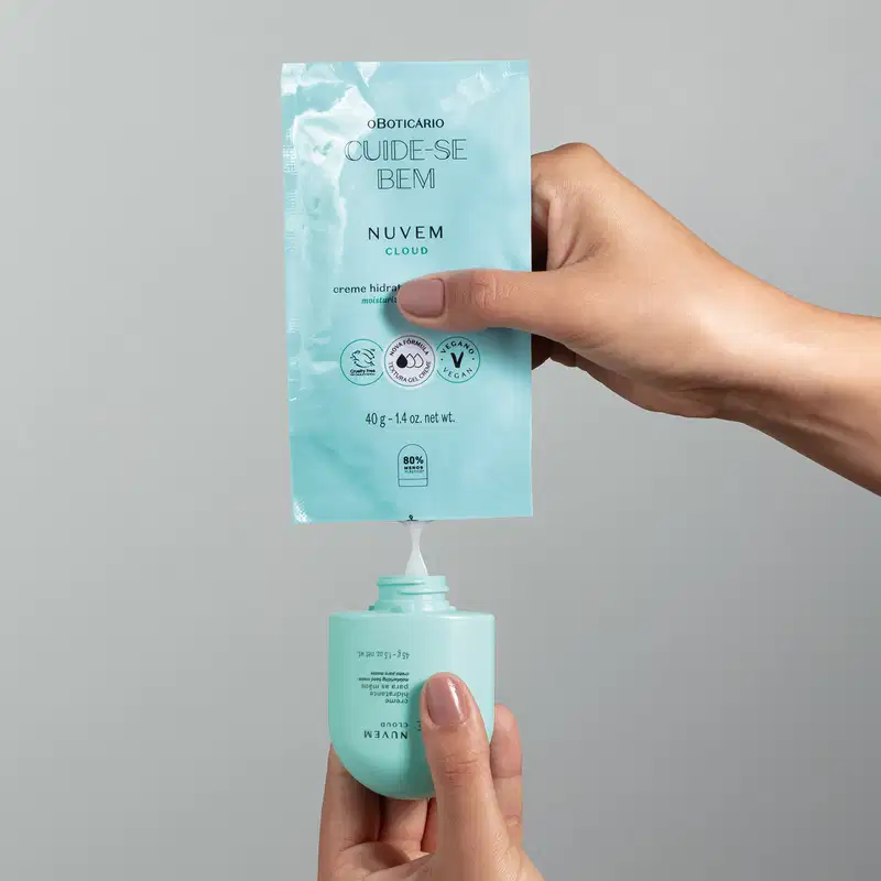 Uma mão feminina segura um sachê azul e espreme um creme branco de creme hidratante para as mãos Cuide-se Bem Nuvem em um frasco original do produto sobre fundo cinza.