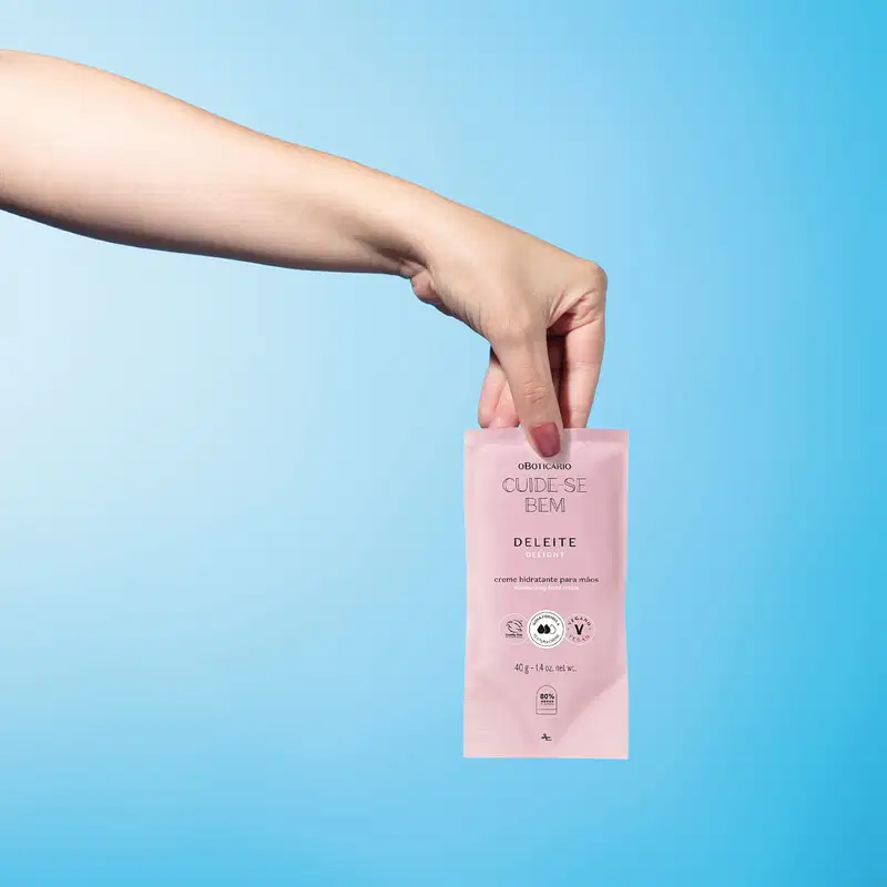 Mão segurando, com a ponta dos dedos, um refil de creme hidratante para as mãos da marca Cuide-se Bem Deleite em sachê na cor rosa com letras pretas. O fundo da imagem é azul vibrante.